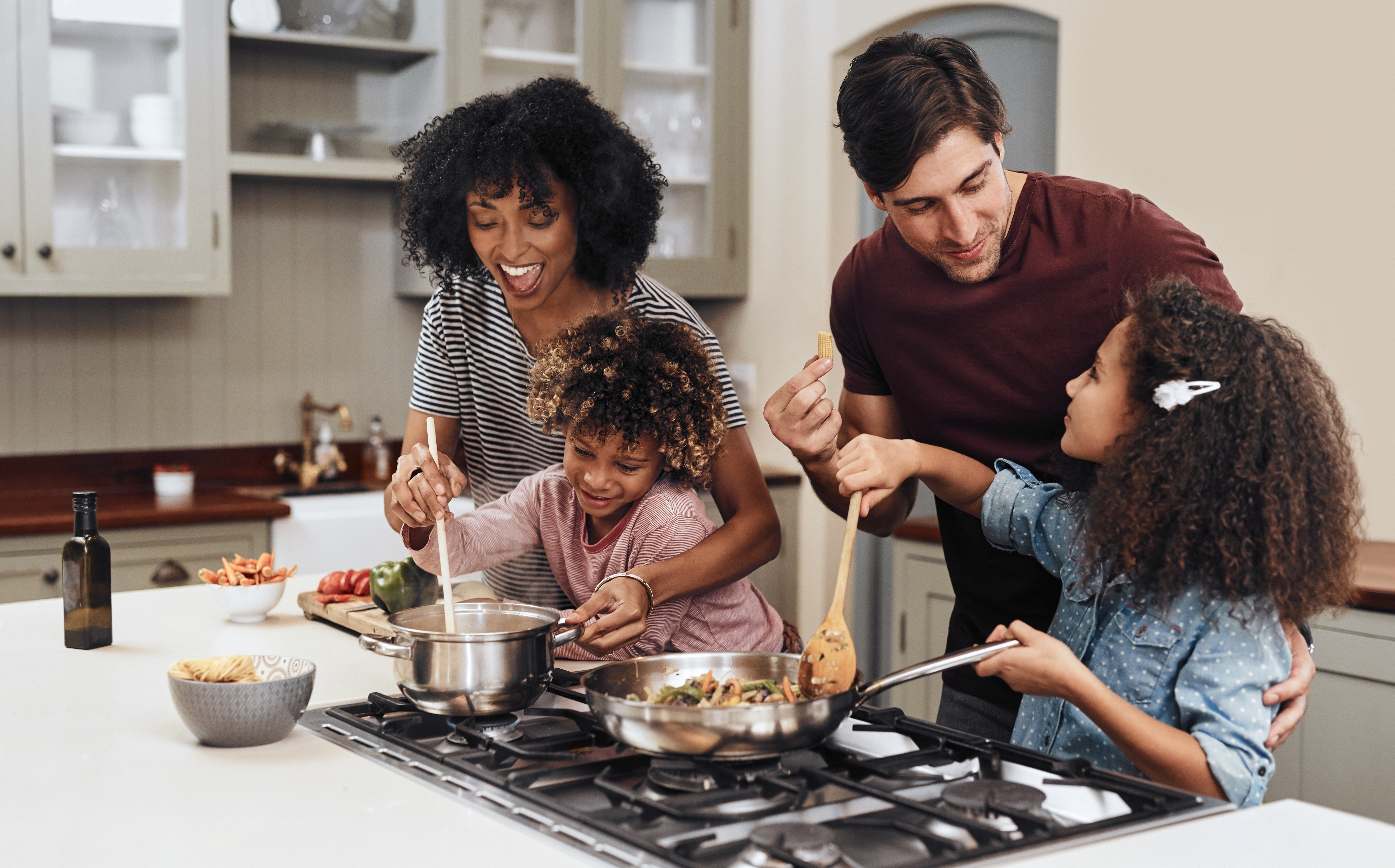 Family of four cooking a meal with kids. Mom and dad are helping their two children saute vegetables in pans on the stove  