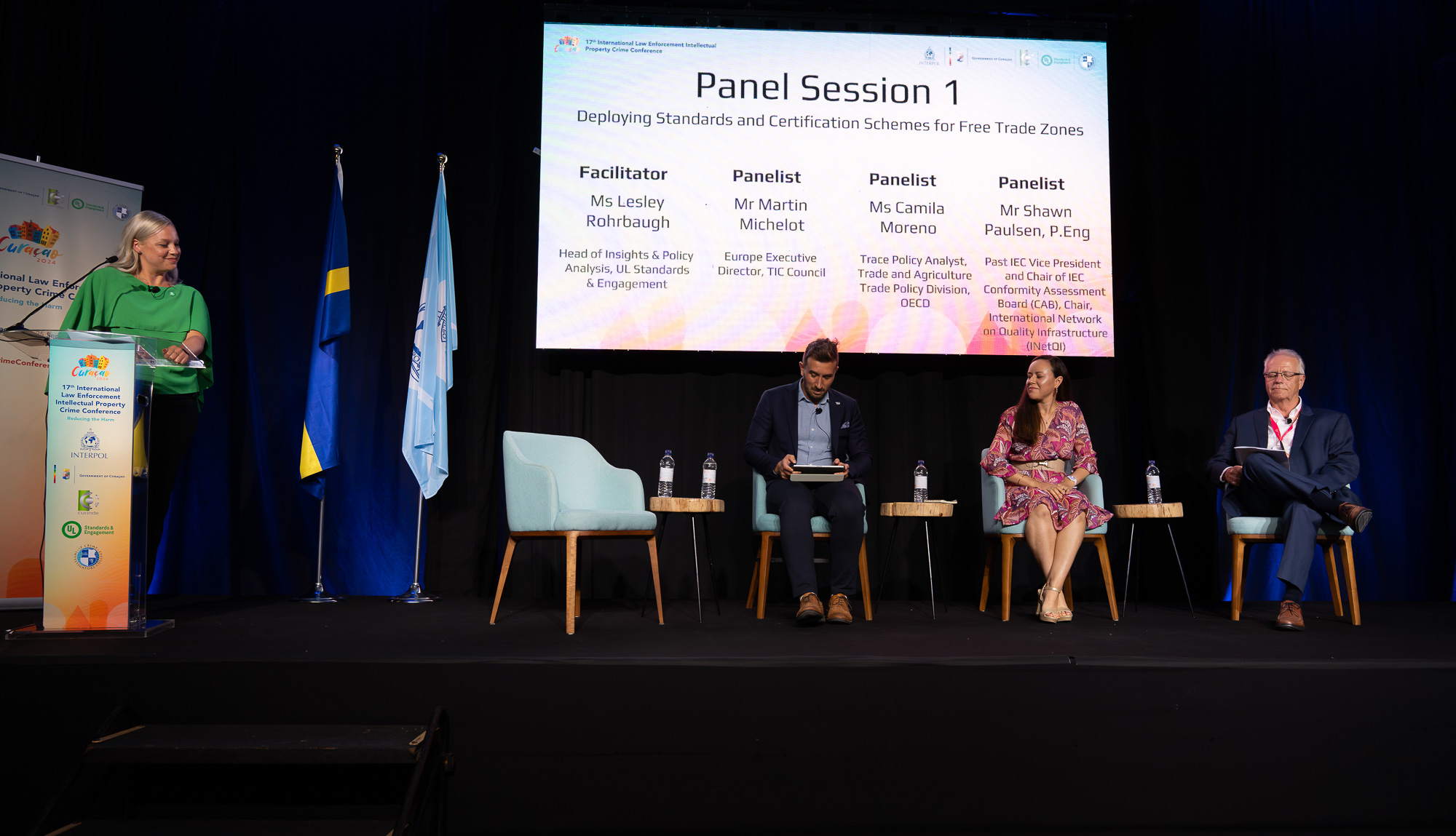 Lesley Rohrbaugh standing at a podium and speaking to panelists at the 2024 International Law Enforcement Intellectual Property Crime Conference in Willemstad, Curaçao