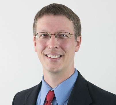 A picture of Dr. J Bern Jordan. A man with glasses wearing a dark blue suit with a light blue shirt and dark red tie.