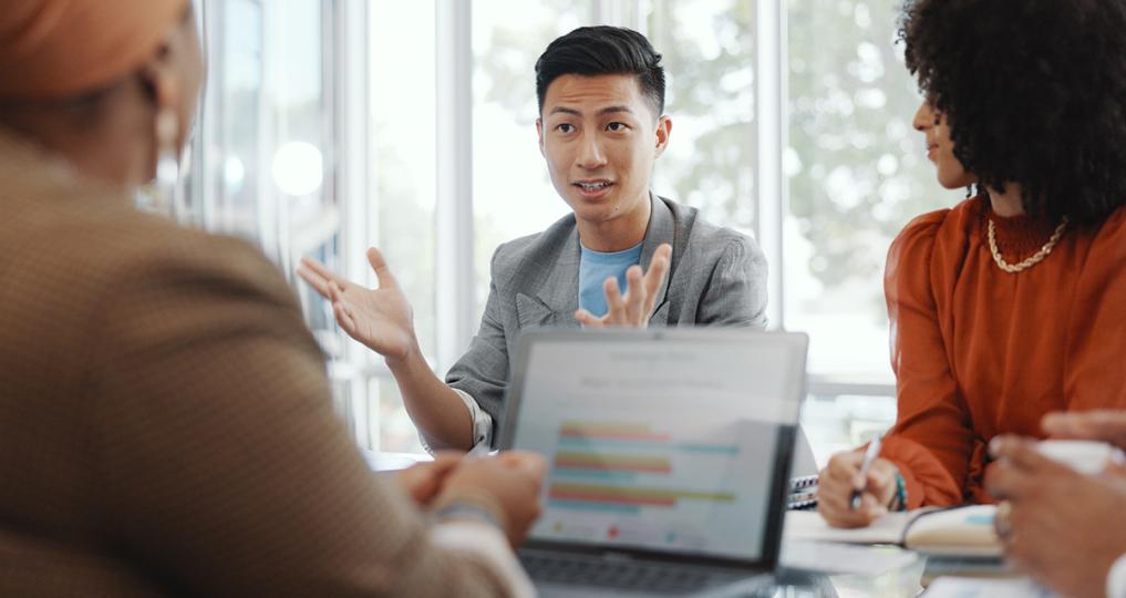 An Asian man in a gray jacket speaking during meeting. A laptop with graphs and data is visible in the foreground.