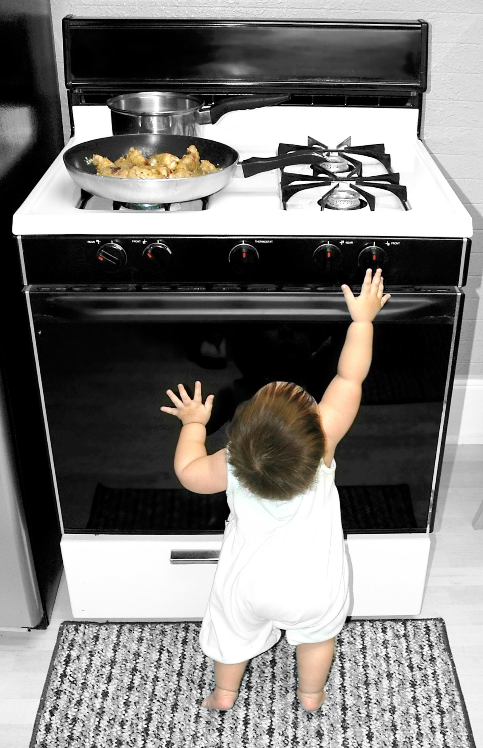 Baby reaching for oven knobs with pan of hot food on range top