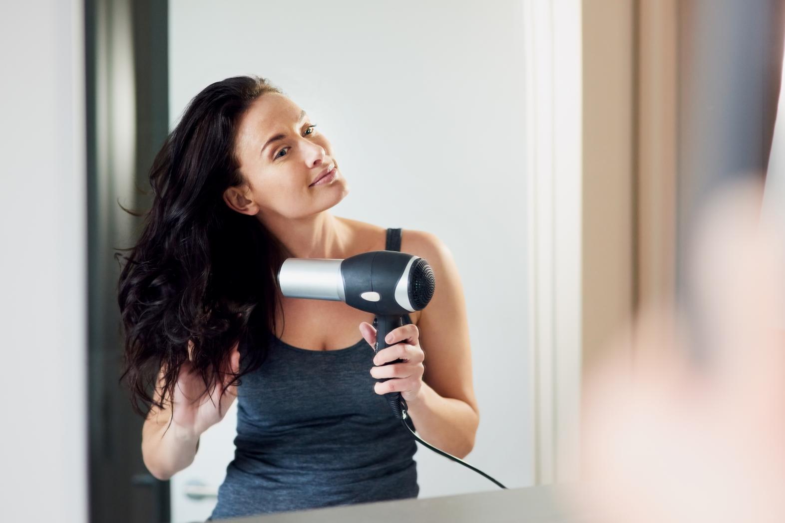 Woman using an electric hair dryer to blow dry her hair in the bathroom mirror