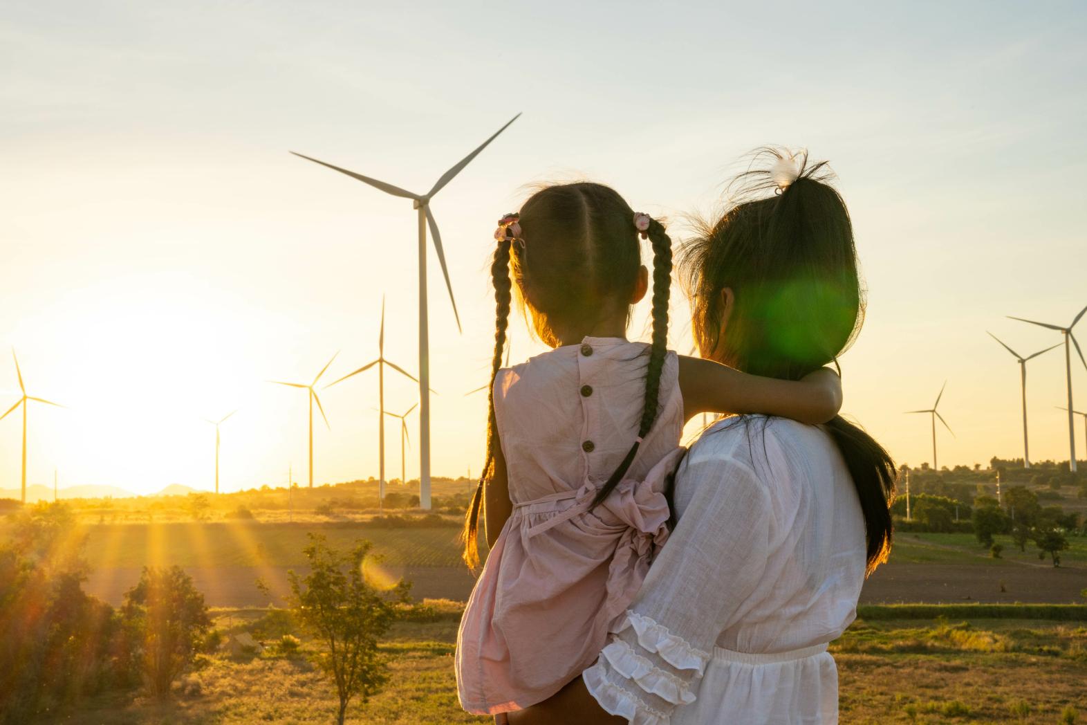 A mother and daughter gaze upon a sunset on a landscape dotted with windmills.