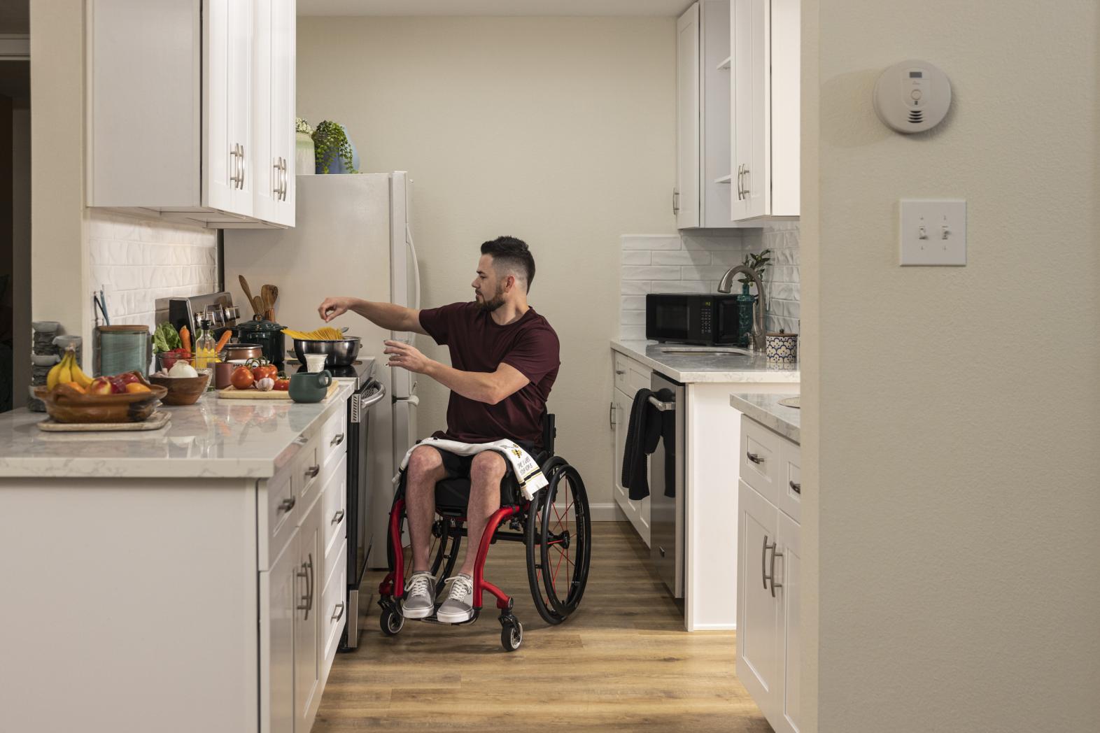 Man in a wheelchair preparing a meal in an apartment kitchen with countertops and appliances at an accessible height