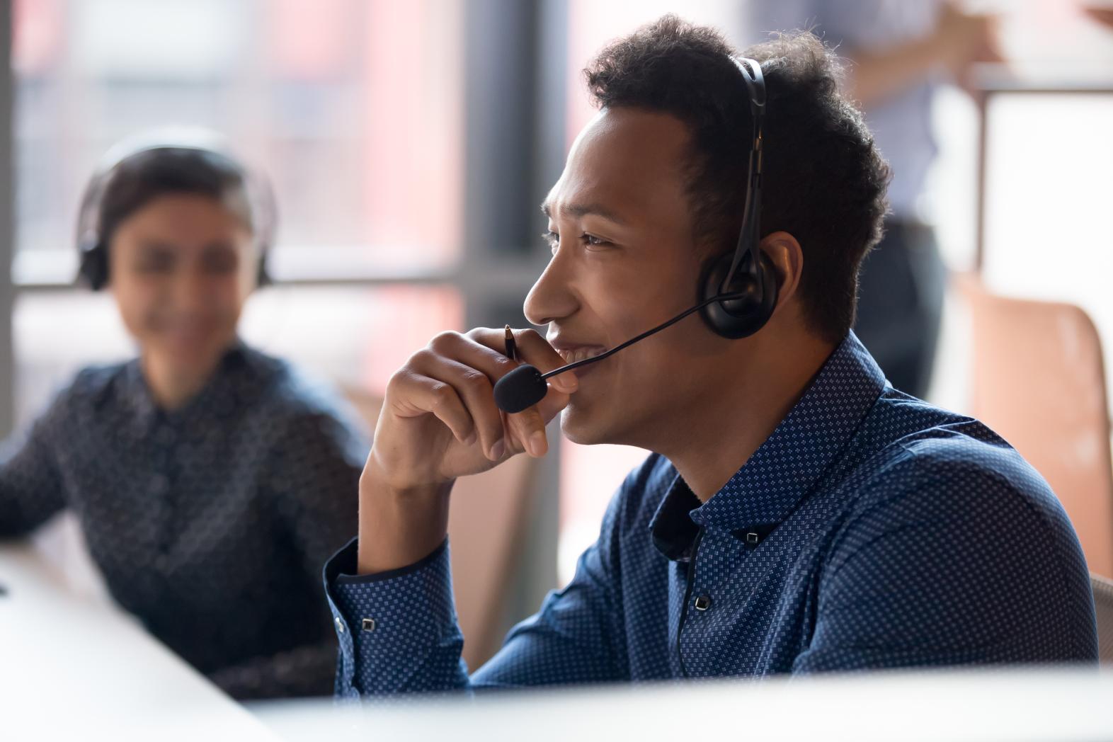 Employees in a call center smiling while working. A man is in the foreground wearing a headset. A female co-worker is observing in the background