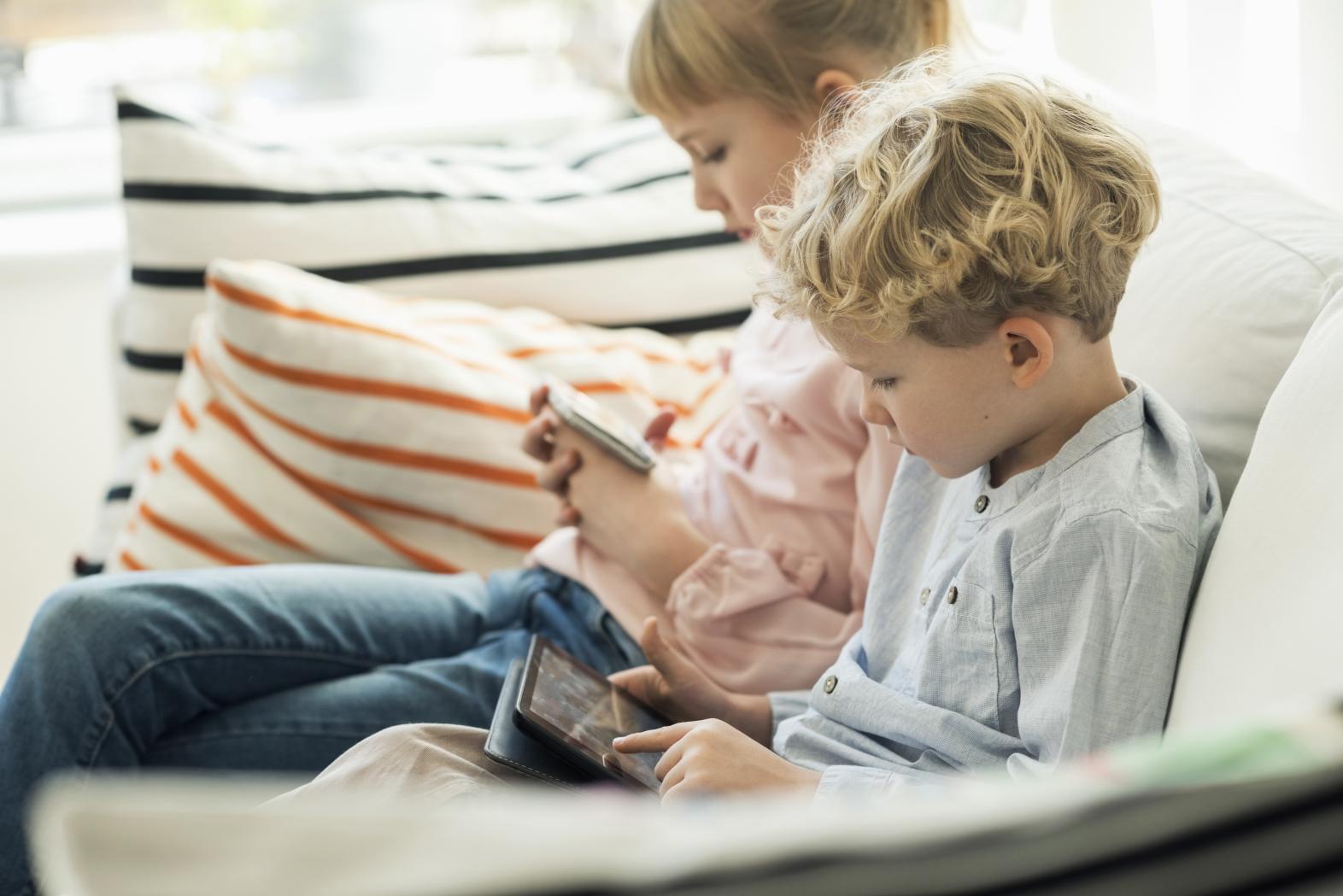 Two children seated on a sofa looking at a tablet and e-reader