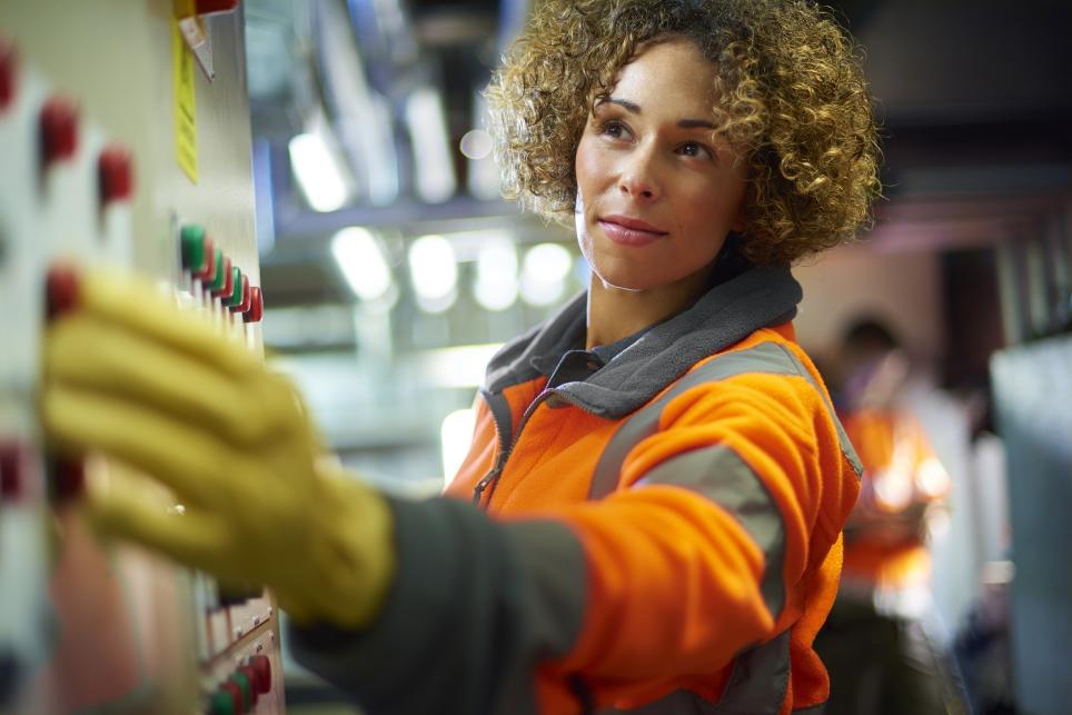 Female engineer in protective gear flips a switch on a panel in an industrial facility