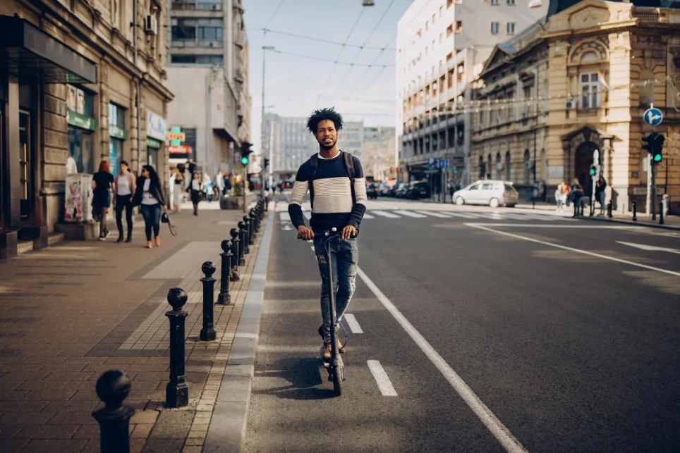 Young African-American man riding an electric scooter on a city street while wearing a backpack. He is smiling and wearing a blue striped sweater.