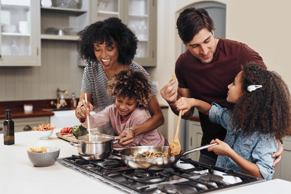 Family of four cooking a meal with kids. Mom and dad are helping their two children saute vegetables in pans on the stove  