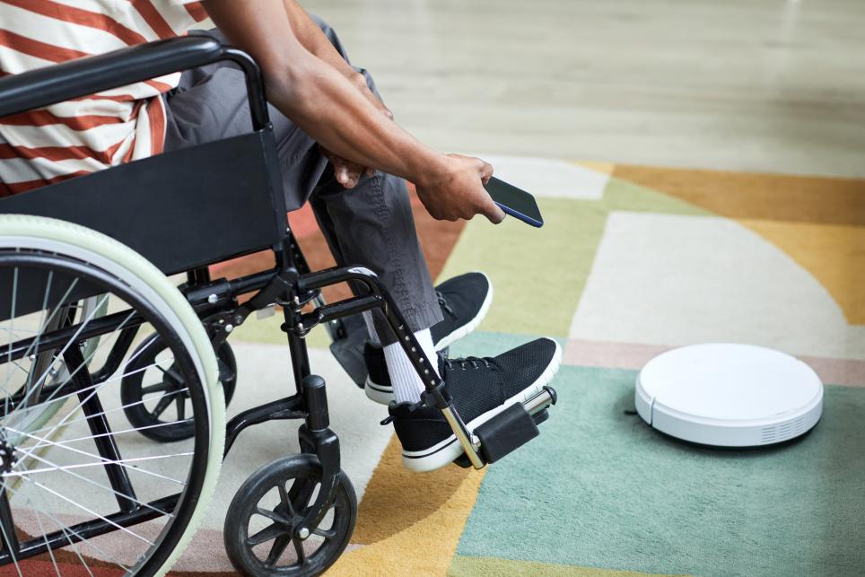 African-american man in wheelchair using smartphone to control a robot vaccuum.