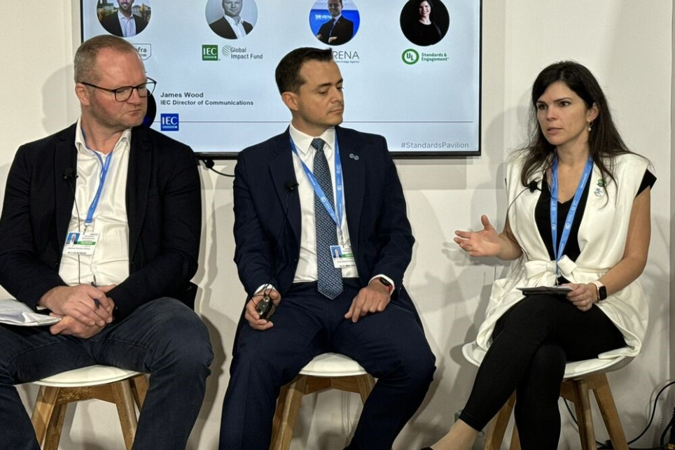 A woman seated on a stage is speaking while two men look on and listen