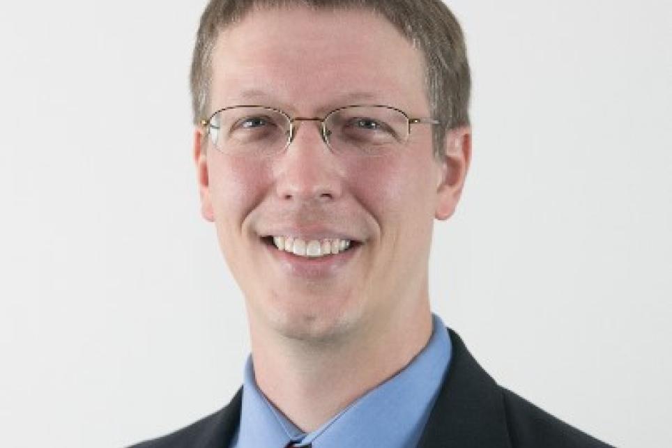 A picture of Dr. J Bern Jordan. A man with glasses wearing a dark blue suit with a light blue shirt and dark red tie.