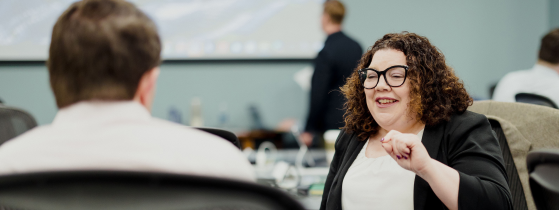 Woman with glasses and curly hair speaking to colleague in meeting