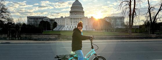 A woman wearing a beanie and smiling riding a bright blue e-bike in front of the US Capitol building.