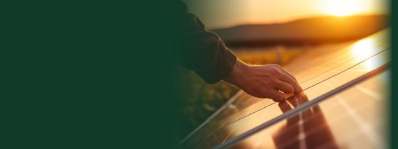 A man's hand rests on a solar panel in the foreground; the sun sets on the horizon in the background.