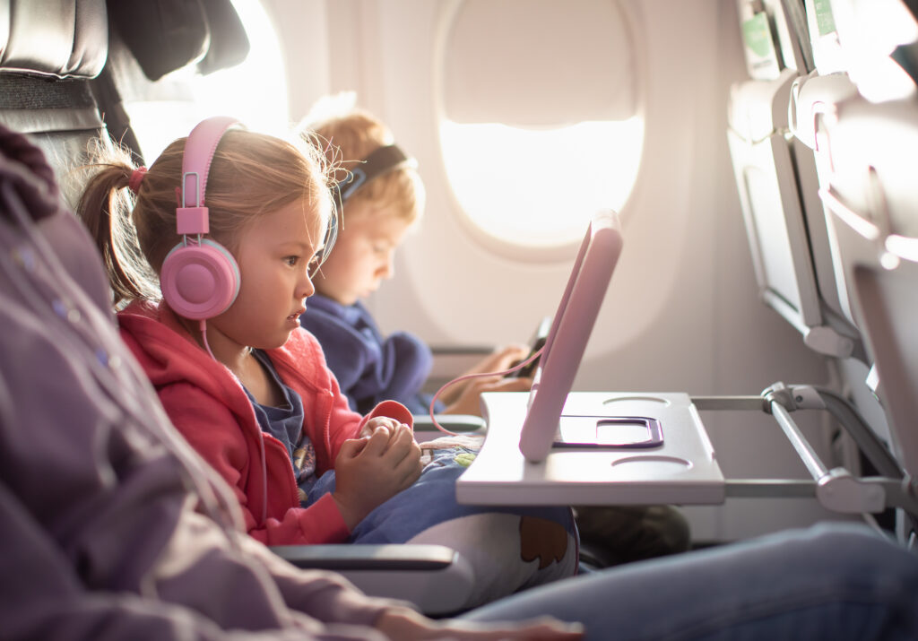 A young girl wearing pink headphones watches a tablet on an airplane tray table, while a boy sits beside her also using a device. Sunlight shines through the airplane window in the background.
