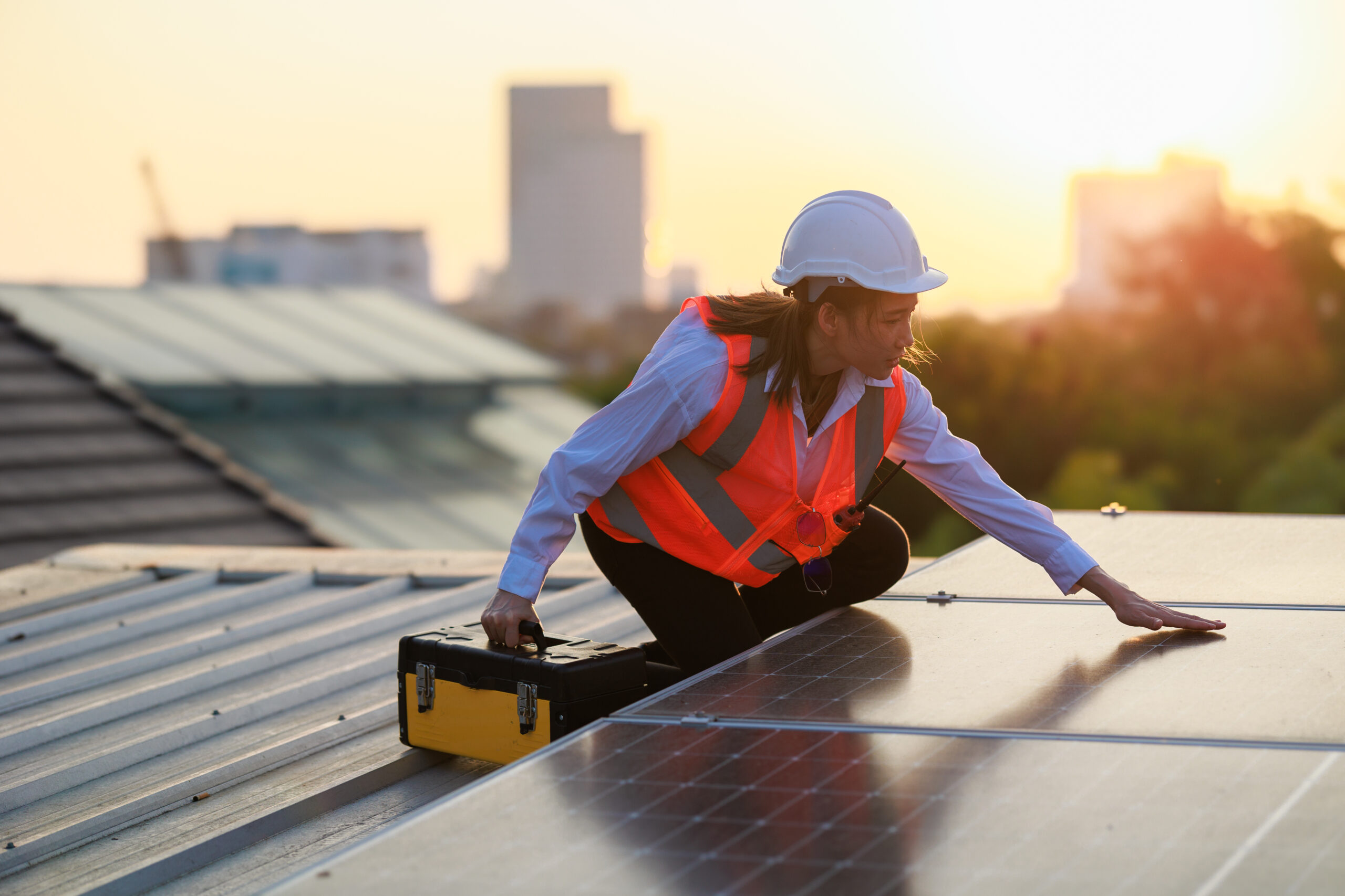 A worker wearing a hard hat and safety vest inspects solar panels on a rooftop at sunset, holding a yellow toolbox with a city skyline in the background.