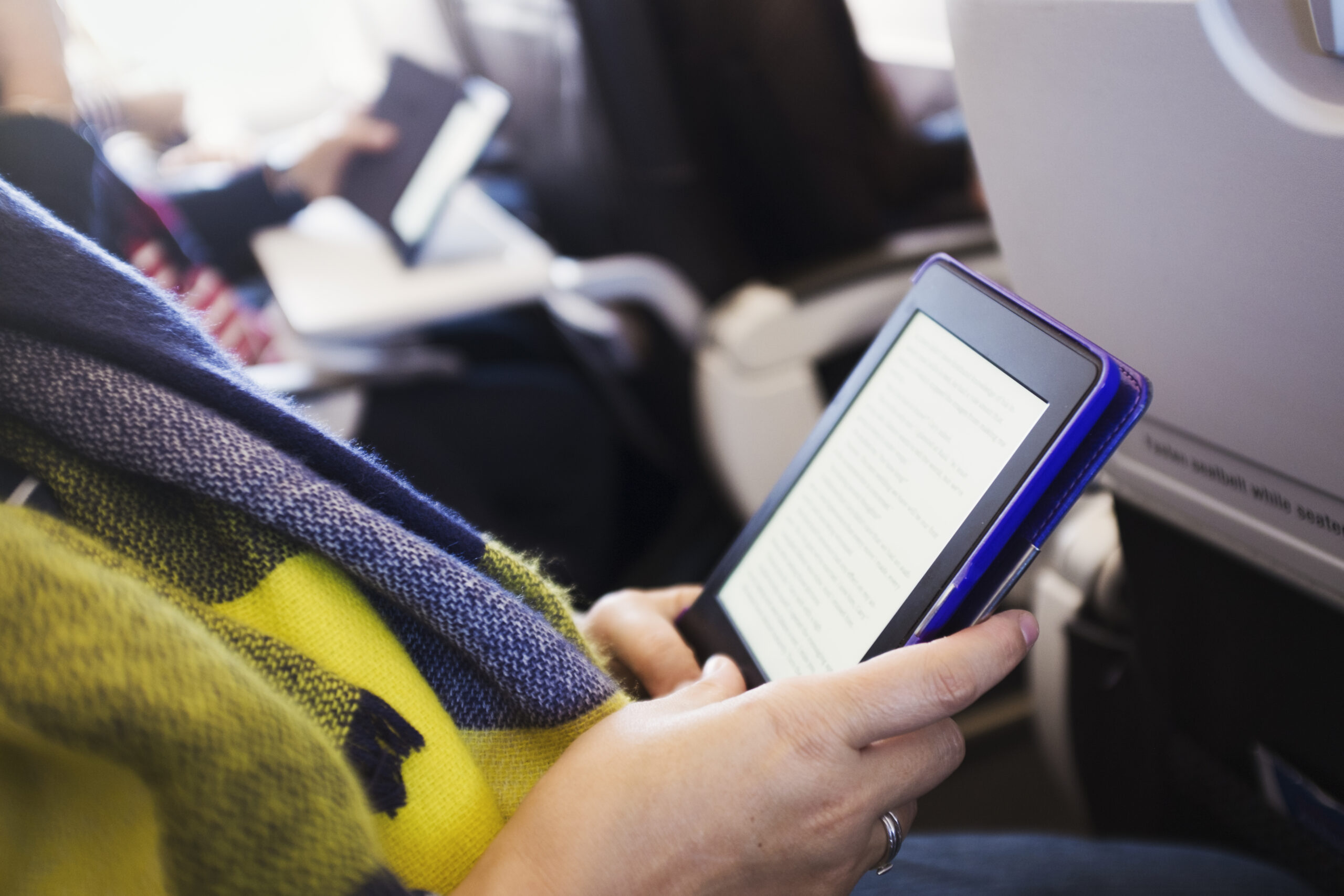 A person wearing a yellow and blue scarf reads an e-book on a tablet while sitting on an airplane, with other passengers and seat trays visible in the background.