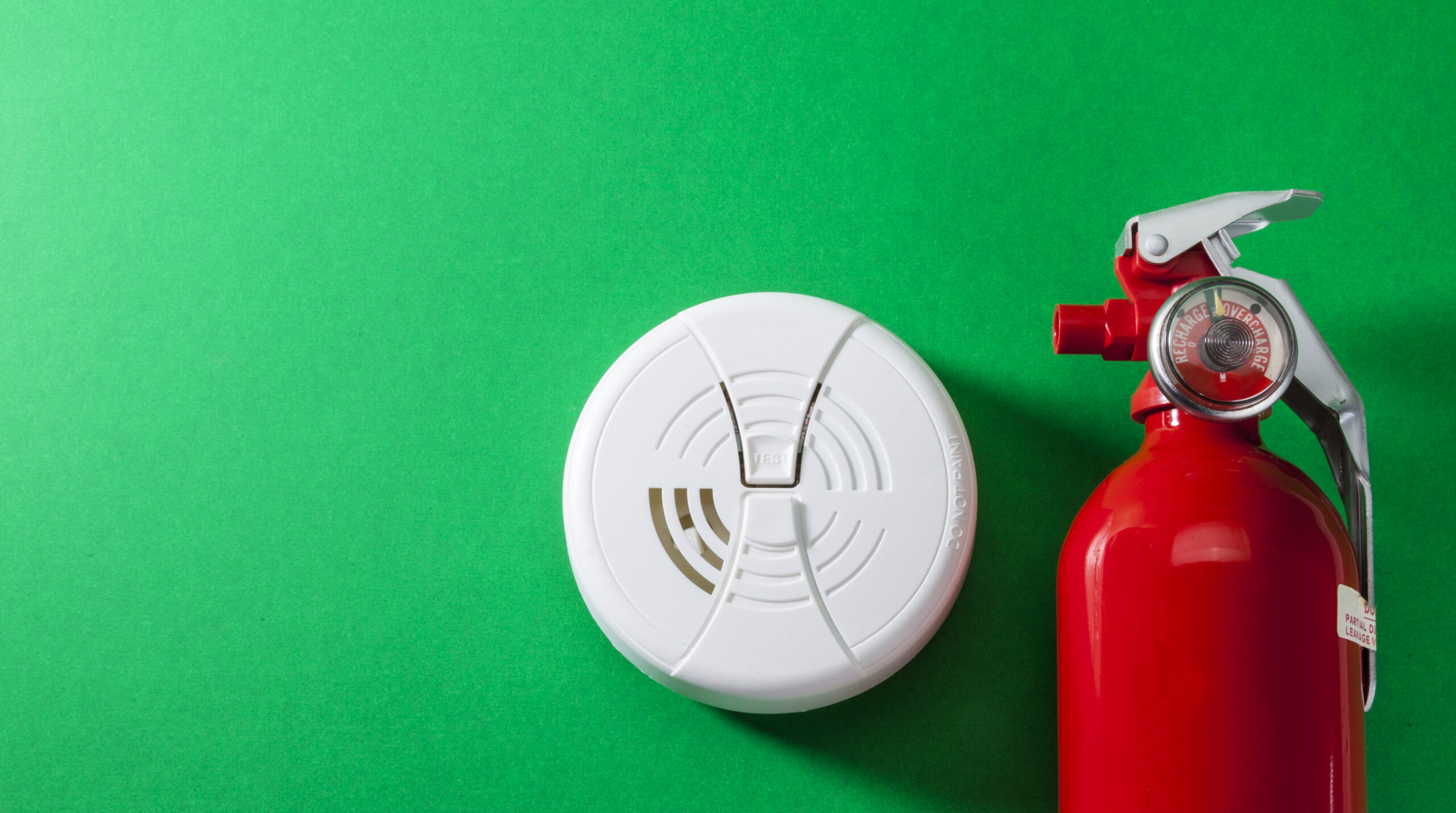 A white smoke detector and a red fire extinguisher are placed side by side on a bright green background.