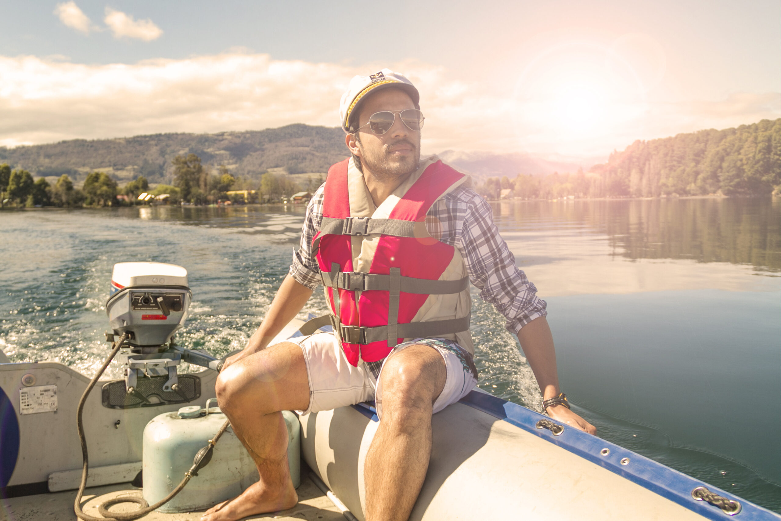 A man wearing sunglasses, a cap, and a life jacket sits in a small motorboat on a calm lake, surrounded by trees and hills under a sunny sky.