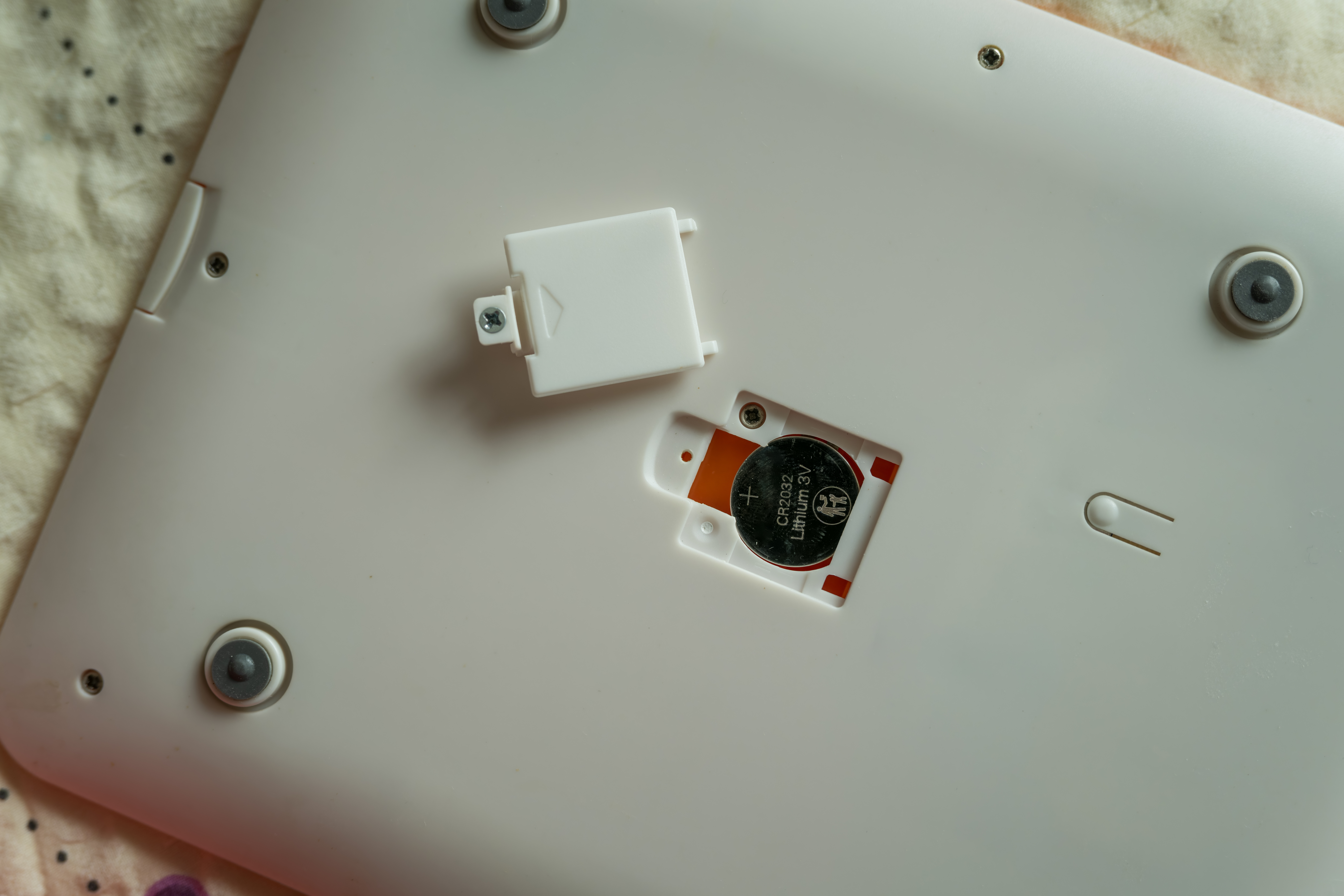 A close-up of the underside of a white electronic device, showing an open battery compartment with a round CR2032 coin cell battery partially exposed and a removed cover next to it.
