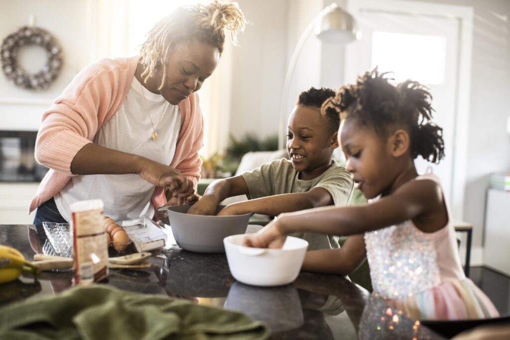 A woman and two children smile and mix ingredients in bowls on a kitchen counter, baking together in a bright, cozy home.
