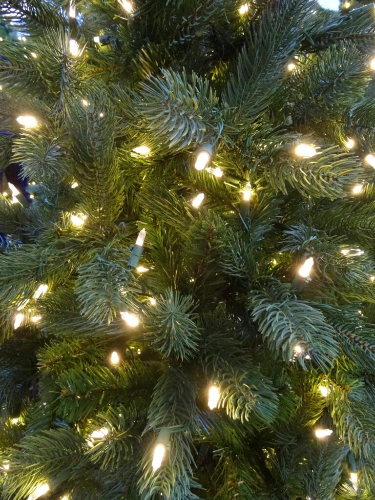Close-up of a green artificial Christmas tree decorated with white string lights, creating a festive and warm holiday atmosphere. The branches are dense and the lights glow softly among the needles.