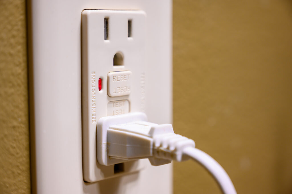 A close-up of a white electrical outlet with a ground fault circuit interrupter (GFCI) showing "reset" and "test" buttons. A white plug is inserted into the lower socket, and a red indicator light is on.