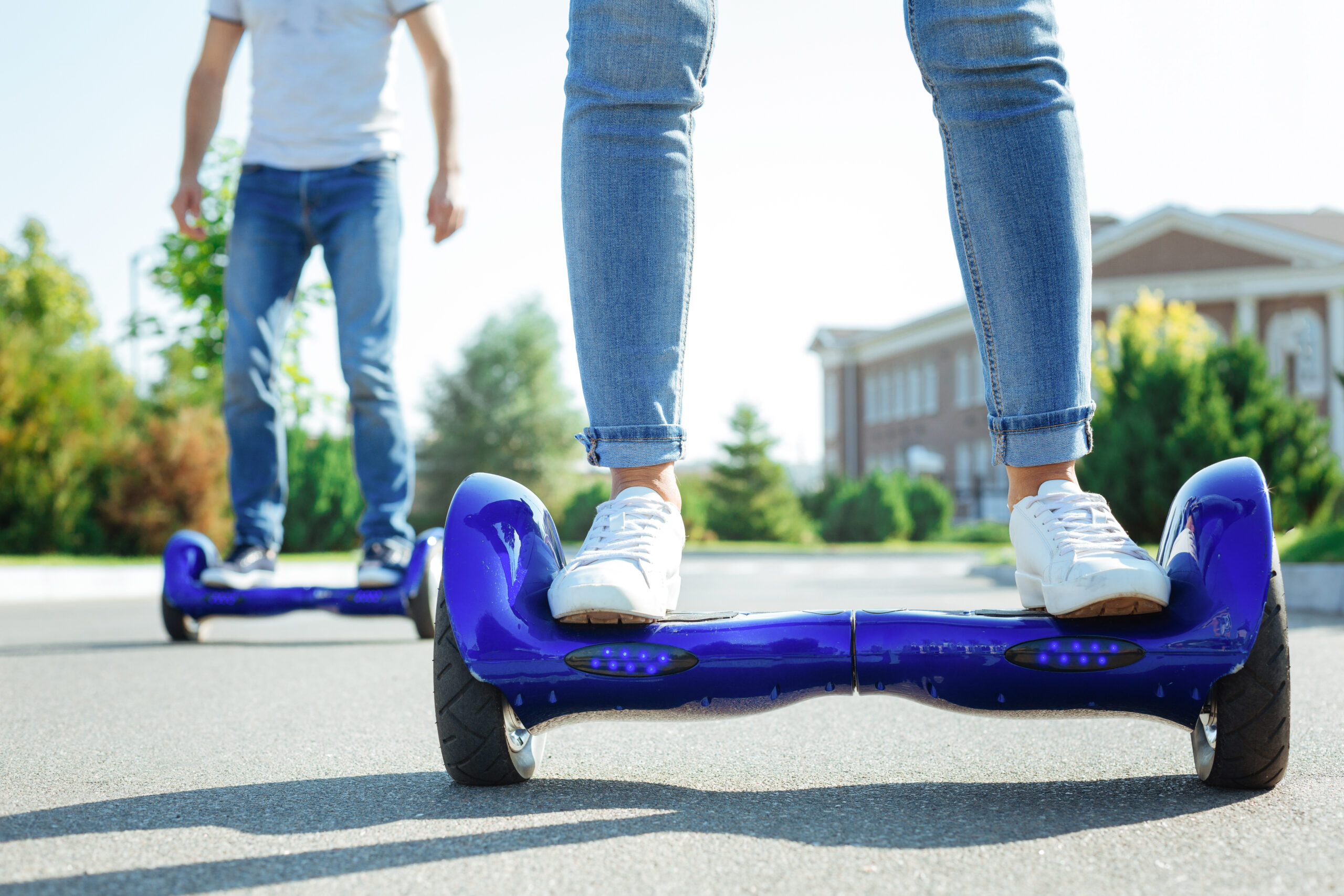 Two people riding blue hoverboards on a street outdoors, with only their legs and feet visible. Both are wearing jeans and white sneakers; trees and a building appear in the background.