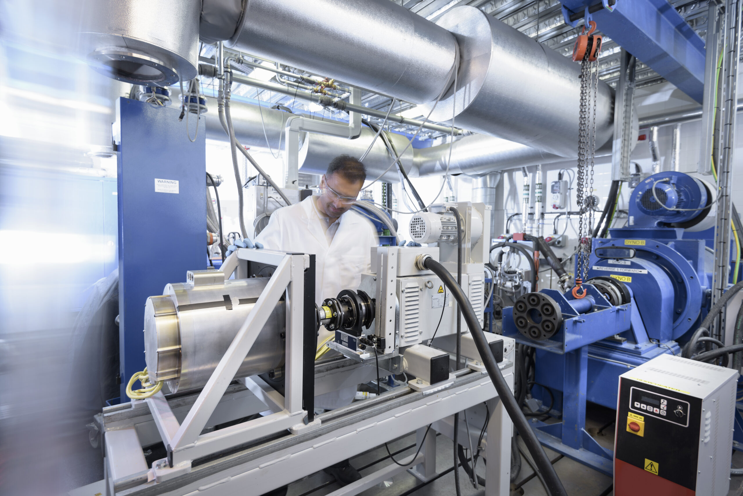 A scientist in a white lab coat and safety glasses operates machinery in an industrial laboratory filled with metal pipes, motors, and various technical equipment.