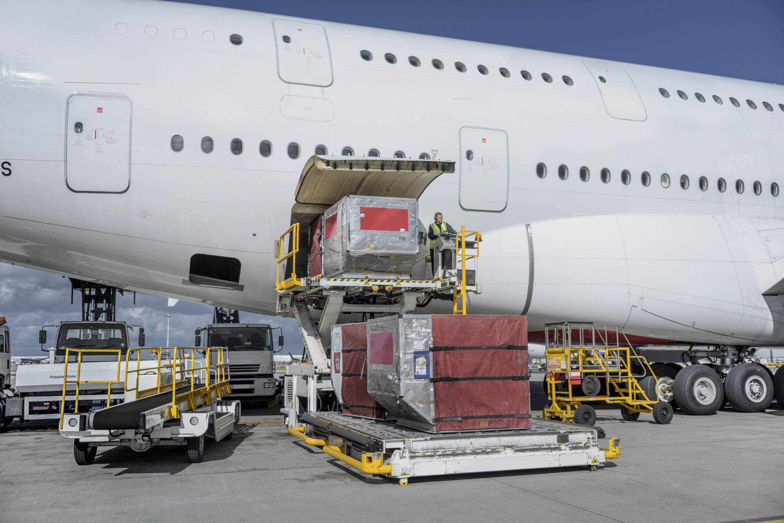 Large red and silver cargo containers are being loaded onto a white airplane using a yellow platform lift on an airport tarmac, with ground support vehicles nearby.