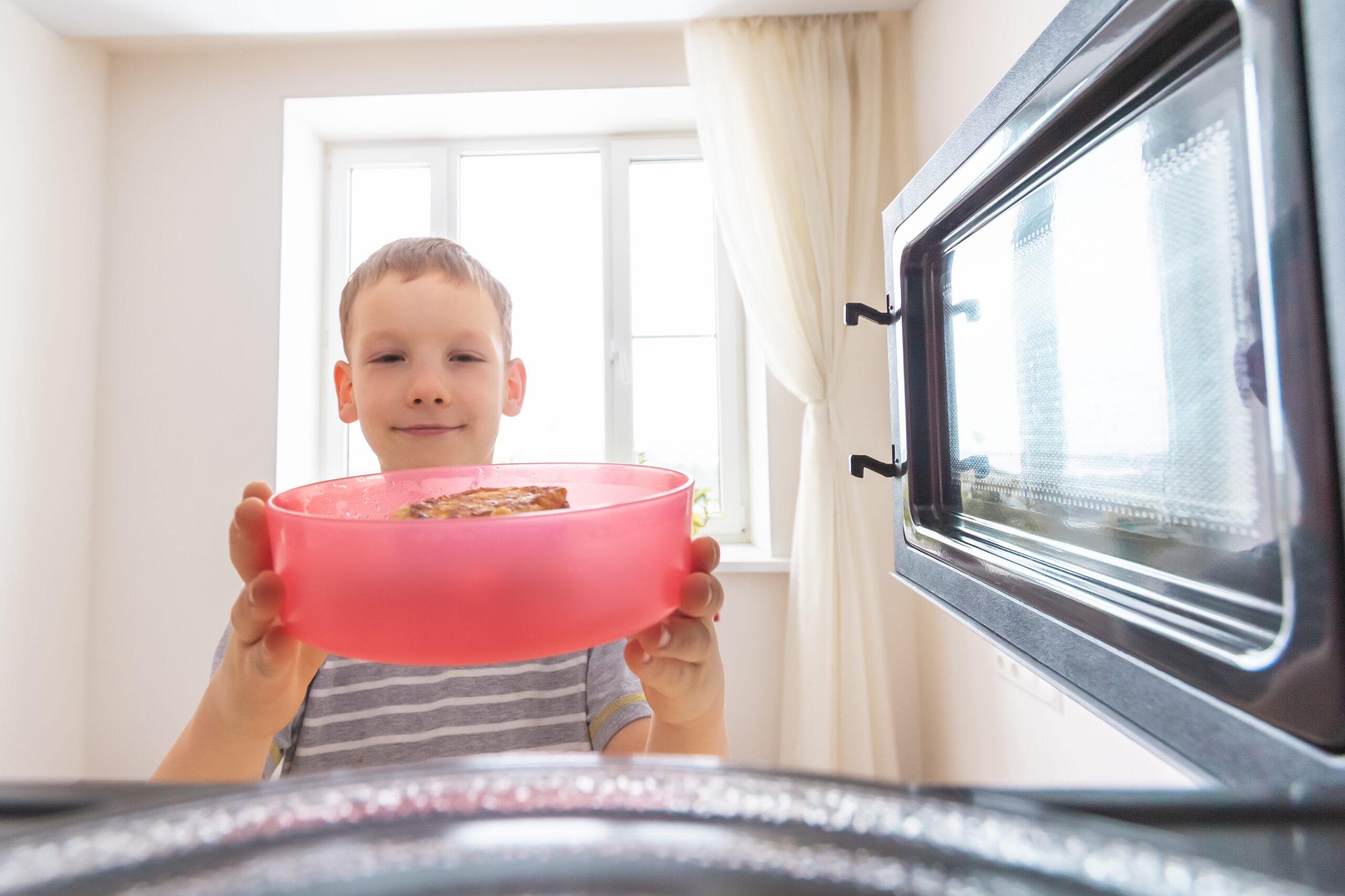 A young boy, smiling, places a red container with food inside a microwave. The photo is taken from inside the microwave, showing the boy and a bright window in the background.