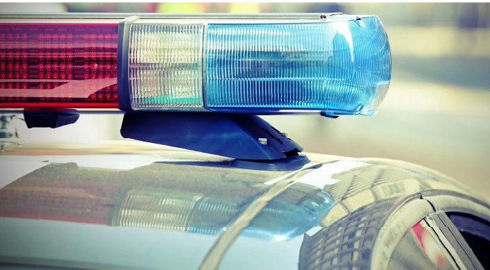 Close-up of red and blue emergency lights on the roof of a police car, with blurred reflections and buildings visible on the car’s surface.