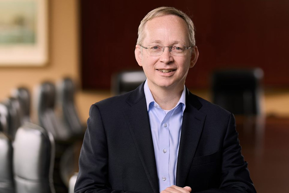 A man in a suit and glasses smiles while standing in a conference room with empty chairs and a blurred background.