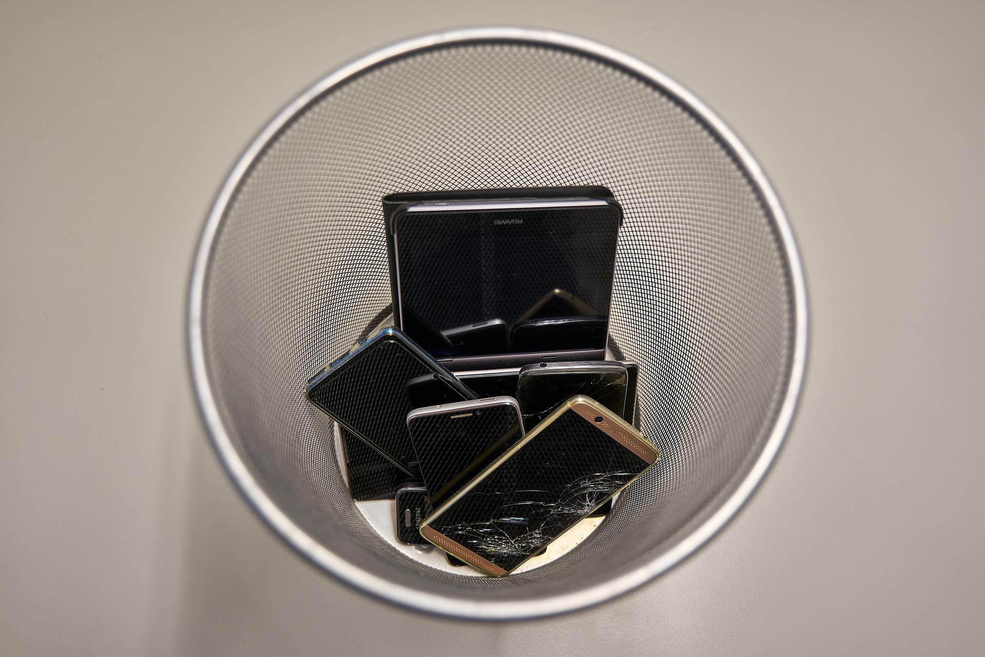 A top-down view of a mesh wastebasket containing several old, broken, and discarded smartphones, some with cracked screens, on a plain, light-colored surface.