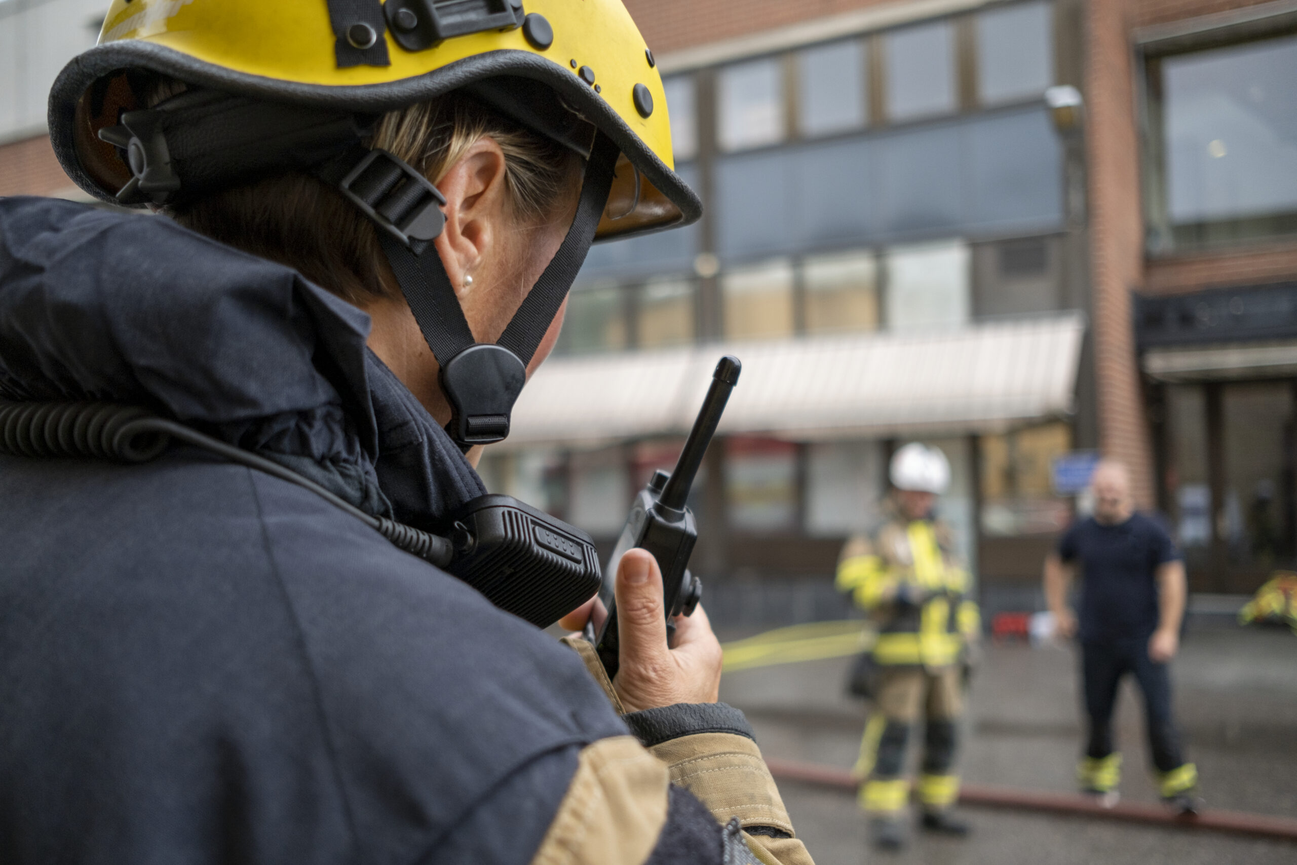 A firefighter in helmet and gear speaks into a walkie-talkie, with two other emergency responders and a building visible in the blurred background.