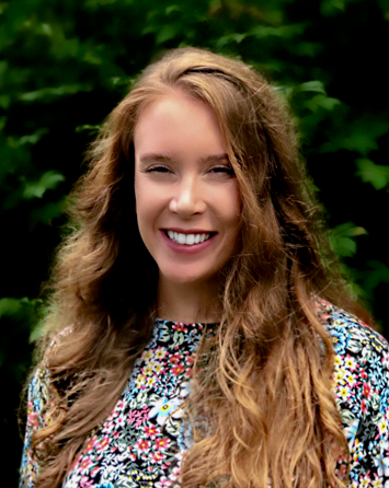 A woman with long, wavy brown hair is smiling outdoors in front of leafy green foliage. She is wearing a colorful floral-patterned top.