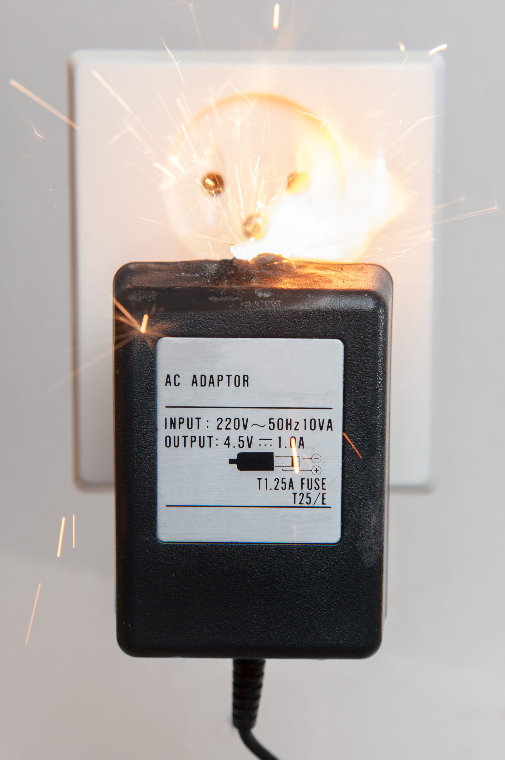 A black AC adapter plugged into a wall socket is sparking, with bright electrical sparks visible around the prongs, indicating a dangerous malfunction or electrical short circuit.