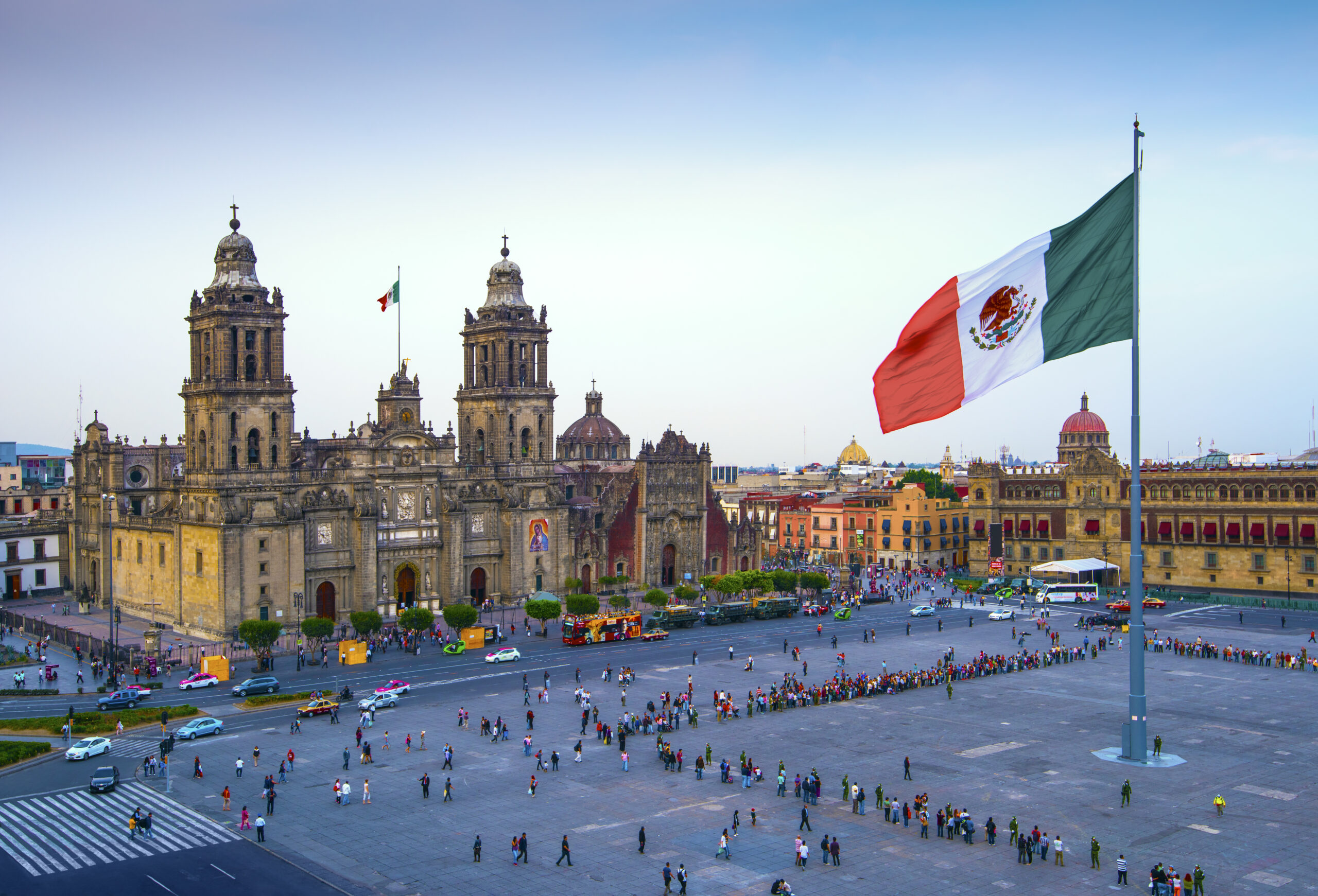 A large Mexican flag waves in the center of Mexico City's Zócalo plaza, surrounded by people. The Metropolitan Cathedral and historic colonial buildings are visible under a clear sky.