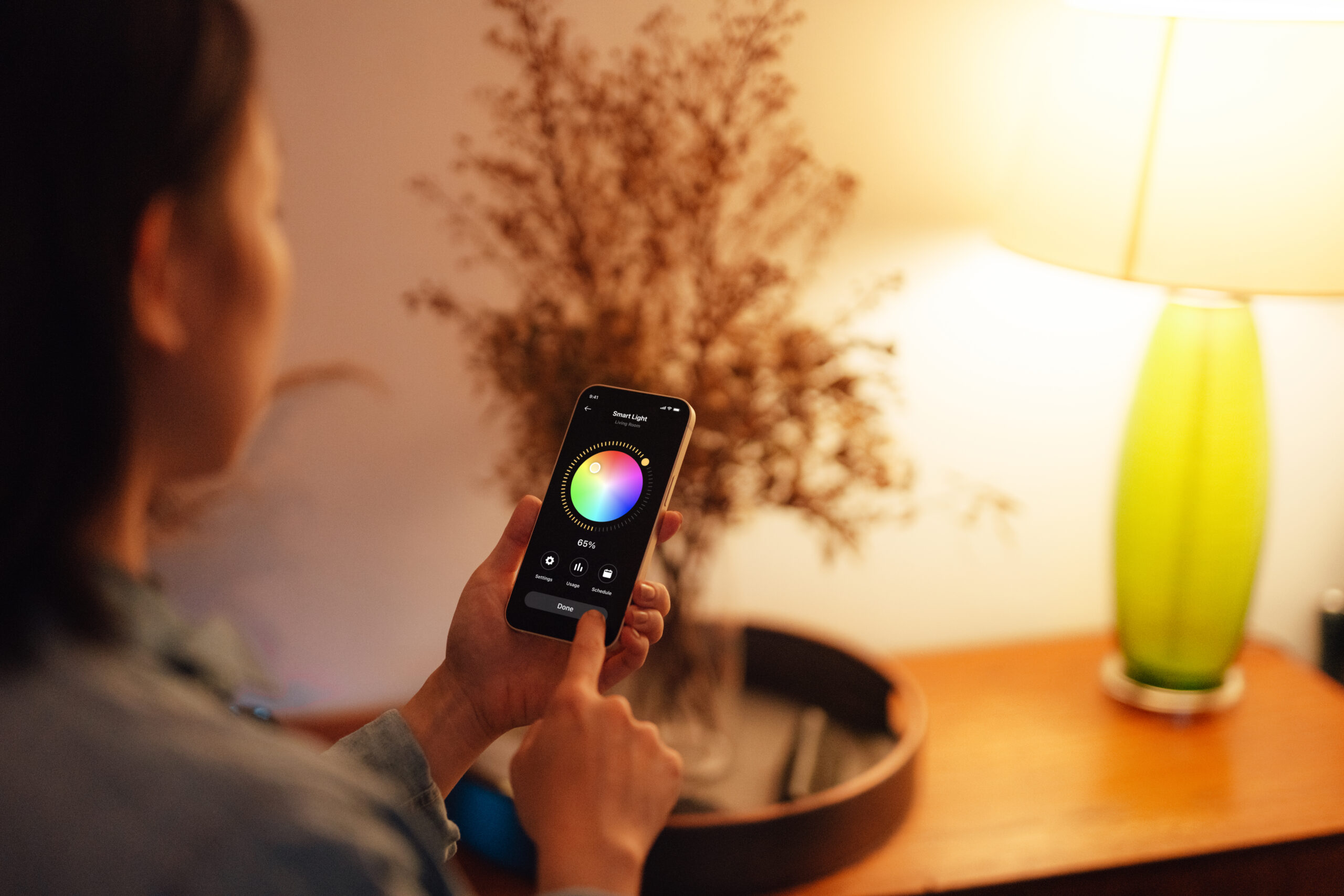 A person adjusts the color and brightness of a smart lamp using a smartphone app. The lamp with a green base is on a wooden table beside dried flowers under warm lighting.