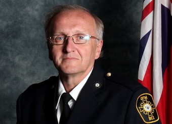 An older man wearing glasses and a dark uniform with a badge on the sleeve poses in front of a flag.