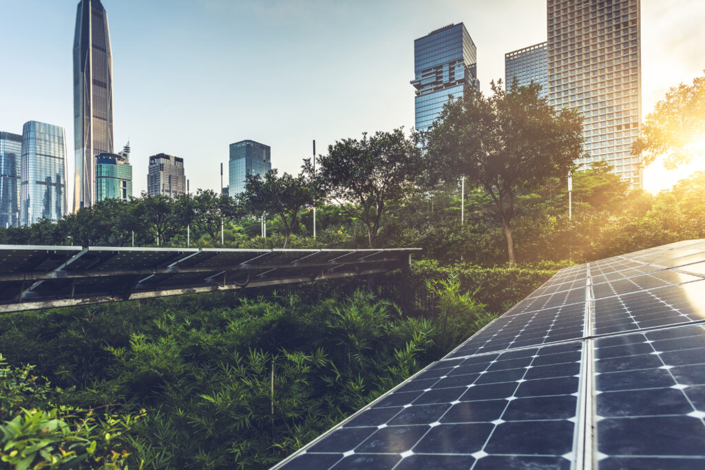 Solar panels in the foreground with modern skyscrapers and tall buildings in the background, surrounded by greenery and trees, with sunlight shining through, representing clean energy in an urban environment.