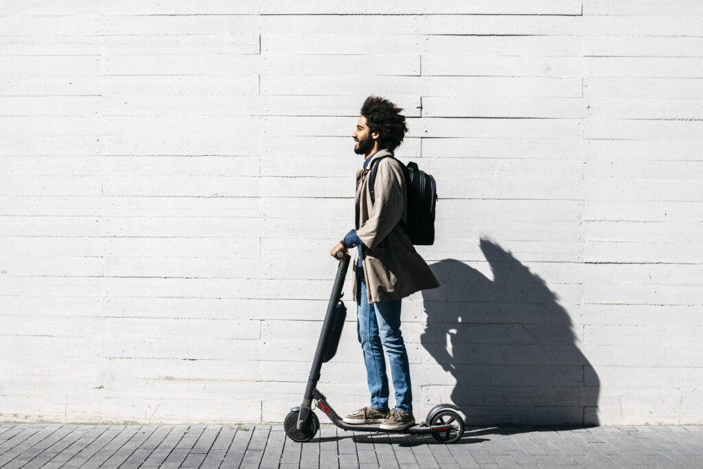 A man with curly hair, wearing glasses, a gray jacket, jeans, and a backpack, rides an electric scooter on a paved sidewalk beside a white concrete wall.
