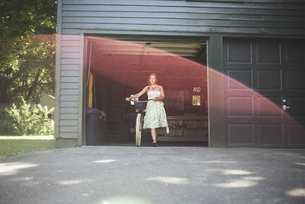 A woman in a white top and patterned skirt stands with a bicycle in the open doorway of a garage. Sunlight creates a soft pink lens flare across the image. Trees and greenery are visible outside.