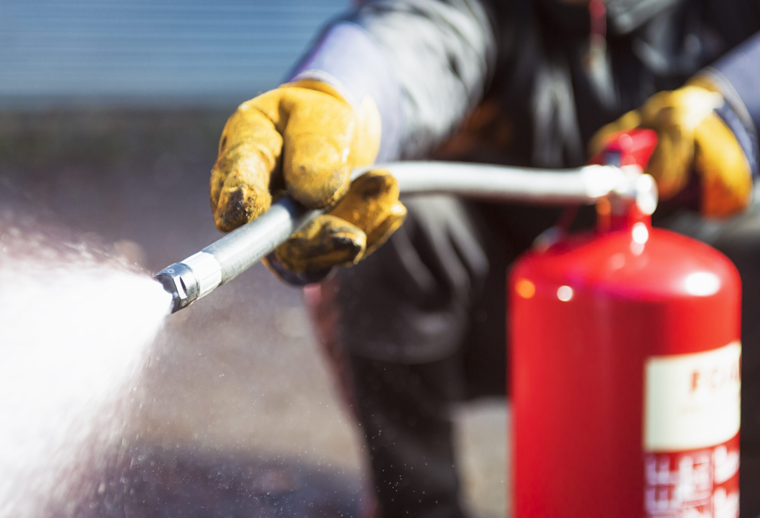 A person wearing yellow gloves uses a red fire extinguisher, with a close-up focus on the nozzle spraying white foam.