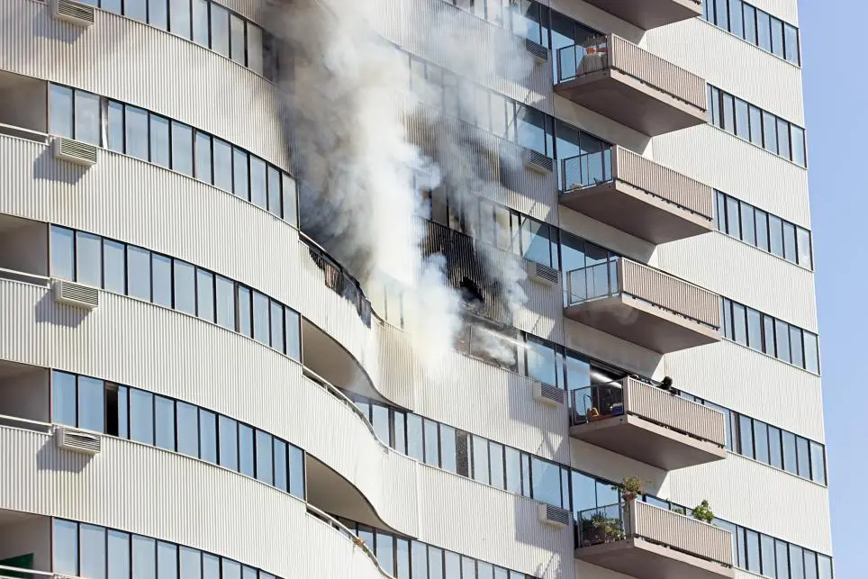 Smoke billows from the windows and balconies of several floors in a high-rise apartment building, indicating an active fire. The exterior of the building is white with curved balcony railings.