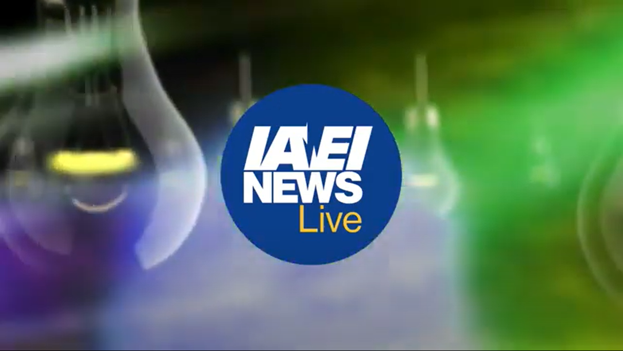 A blue circle with "IAEI NEWS Live" in bold white and yellow text is centered over a blurred background featuring green and purple hues and indistinct hanging lightbulbs.