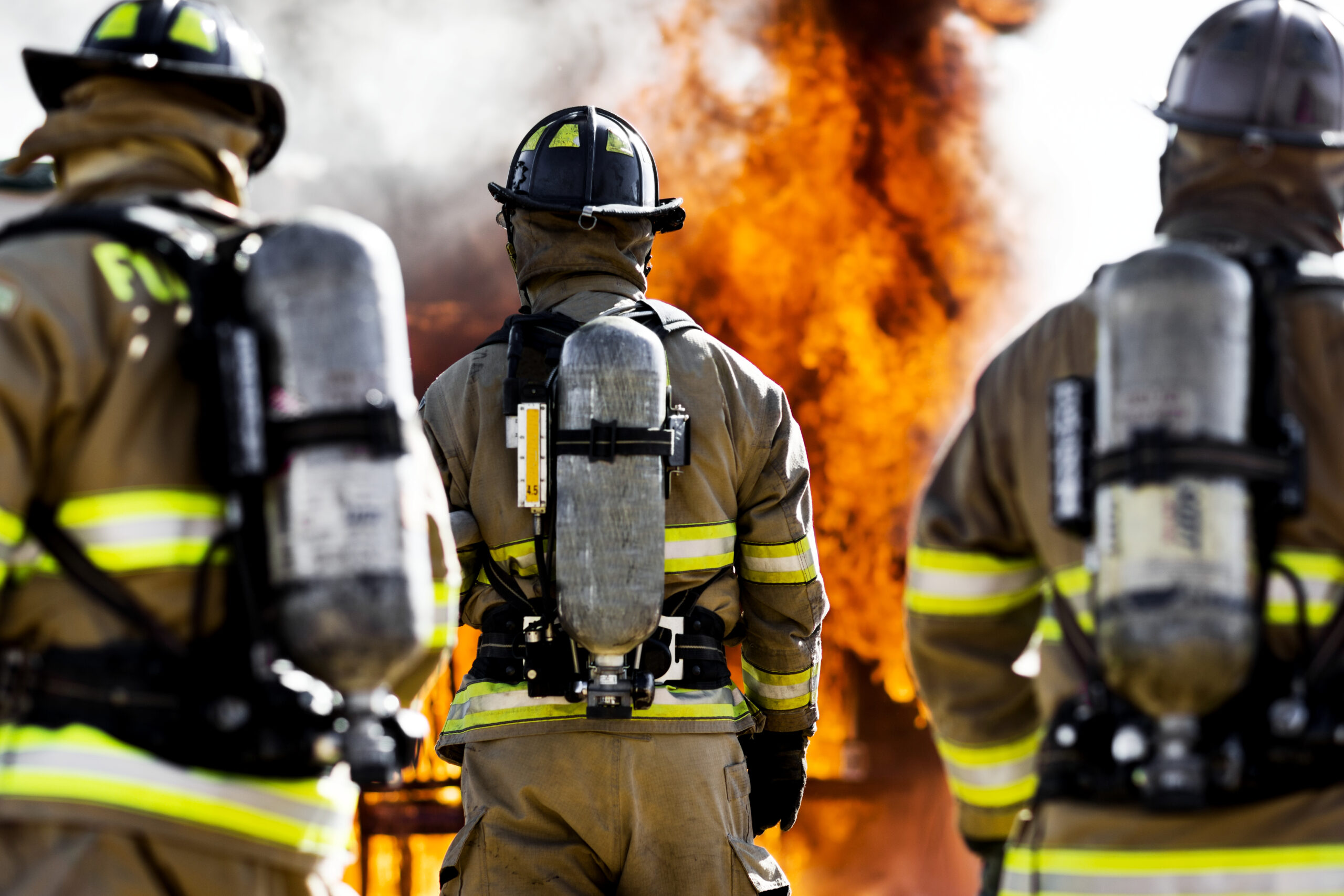 Three firefighters in full gear and oxygen tanks face a large blaze, with bright flames and smoke rising in the background. Their reflective stripes are visible as they prepare to tackle the fire.