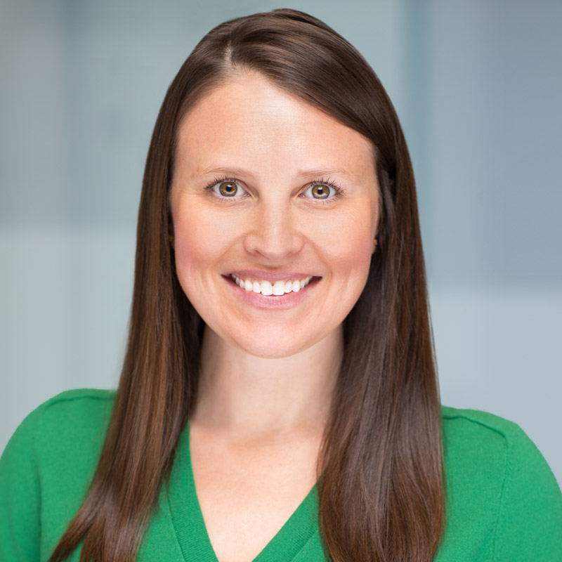 A woman with long straight brown hair and fair skin smiles at the camera. She is wearing a green top and is posed against a blurred light gray background.