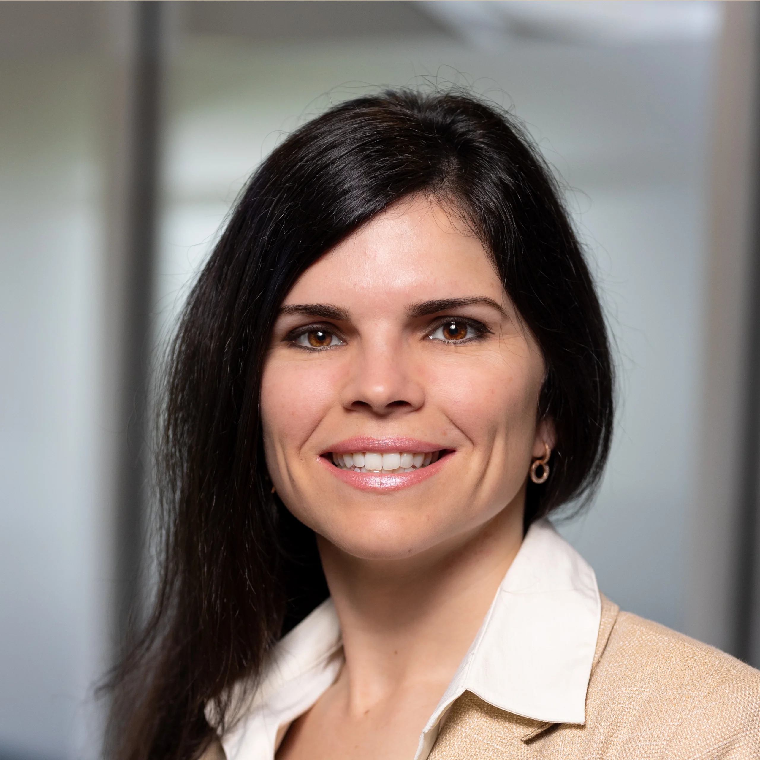 A woman with long dark hair, wearing a beige blazer and white collared shirt, smiles at the camera in an office setting with blurred background.